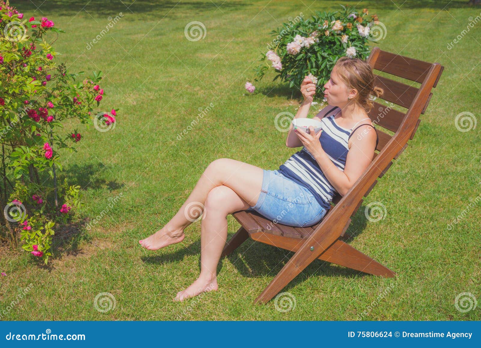 Jovem mulher que relaxa e que come o gelado. Jovem mulher que relaxa na cadeira de jardim e que come o gelado no dia de verão ensolarado