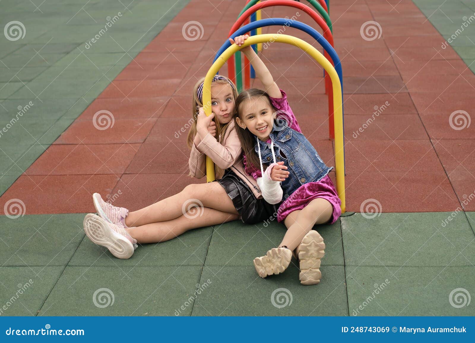 joy little preschool girl with broken arm and cast on her forearm having fun at children`s sports playground with her friend desp