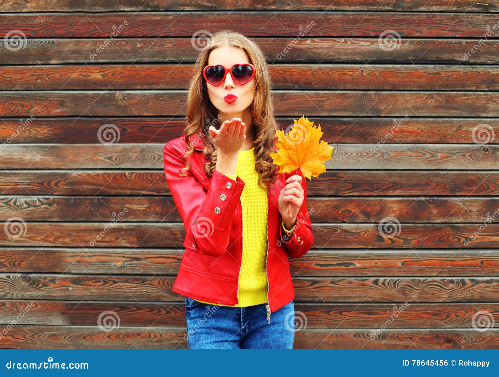 Junge Frau der Mode recht schickt Luft süßen Kuss mit gelben Ahornblättern am Herbsttag über dem hölzernen Hintergrund, der rotes. Junge Frau der Mode recht schickt Luft süßen Kuss mit gelben Ahornblättern am Herbsttag über dem hölzernen Hintergrund, der eine rote Lederjacke trägt