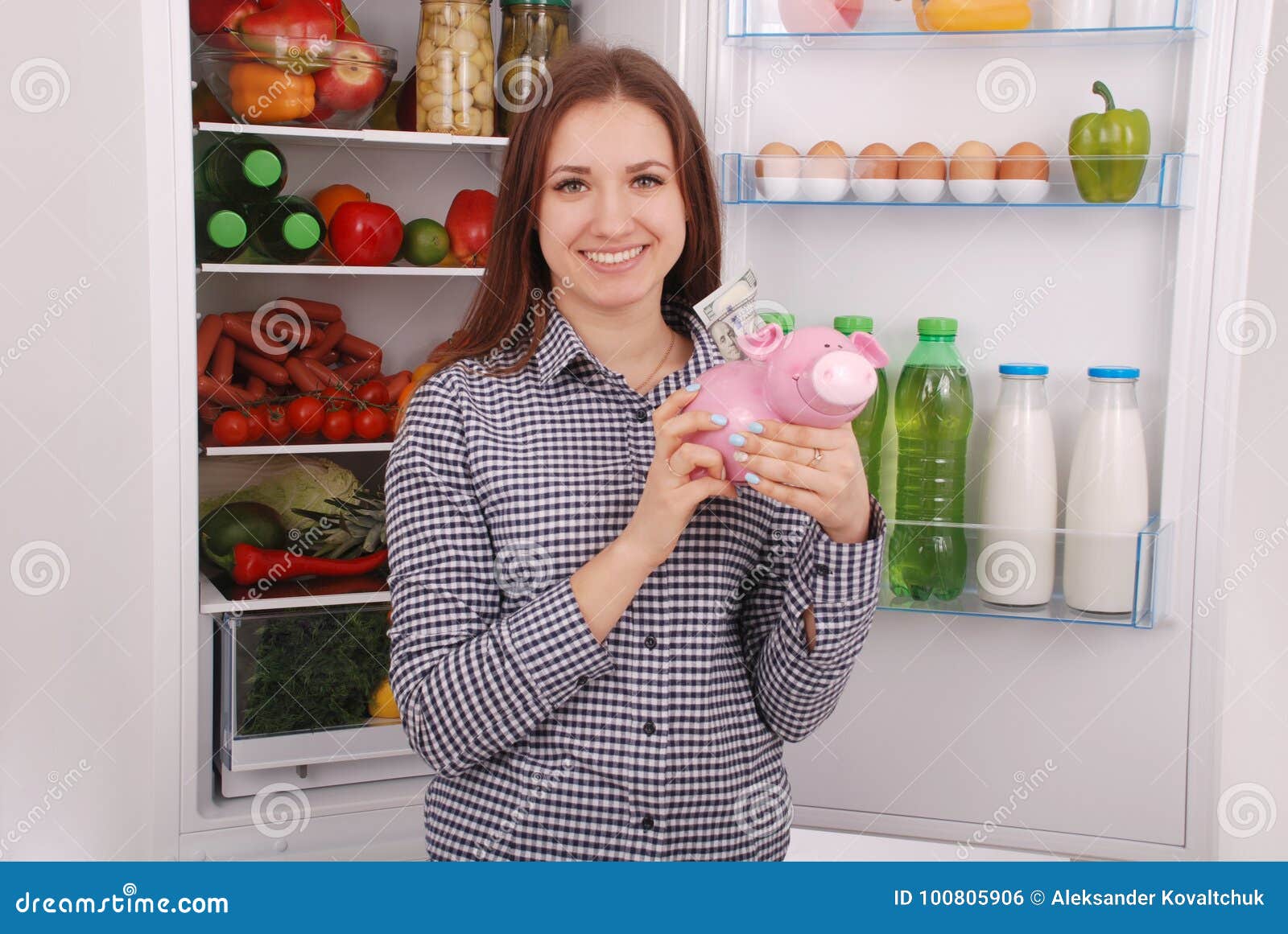 Junge schöne Frau, die mit Geldkasten der piggy Bank steht. Junge Schönheit, die mit Sparschweingeldkasten auf dem Kühlschrankhintergrund steht