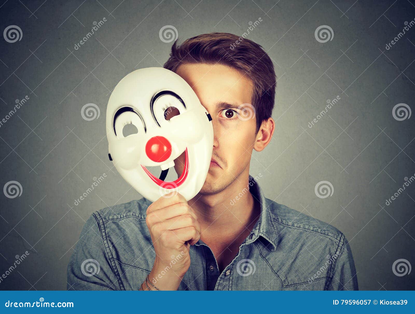 Junger trauriger Mann, der hinter glücklicher Clownmaske sich versteckt. Junger trauriger Mann, der hinter glücklicher Clownmaske auf grauem Wandhintergrund sich versteckt Menschliche Gefühle