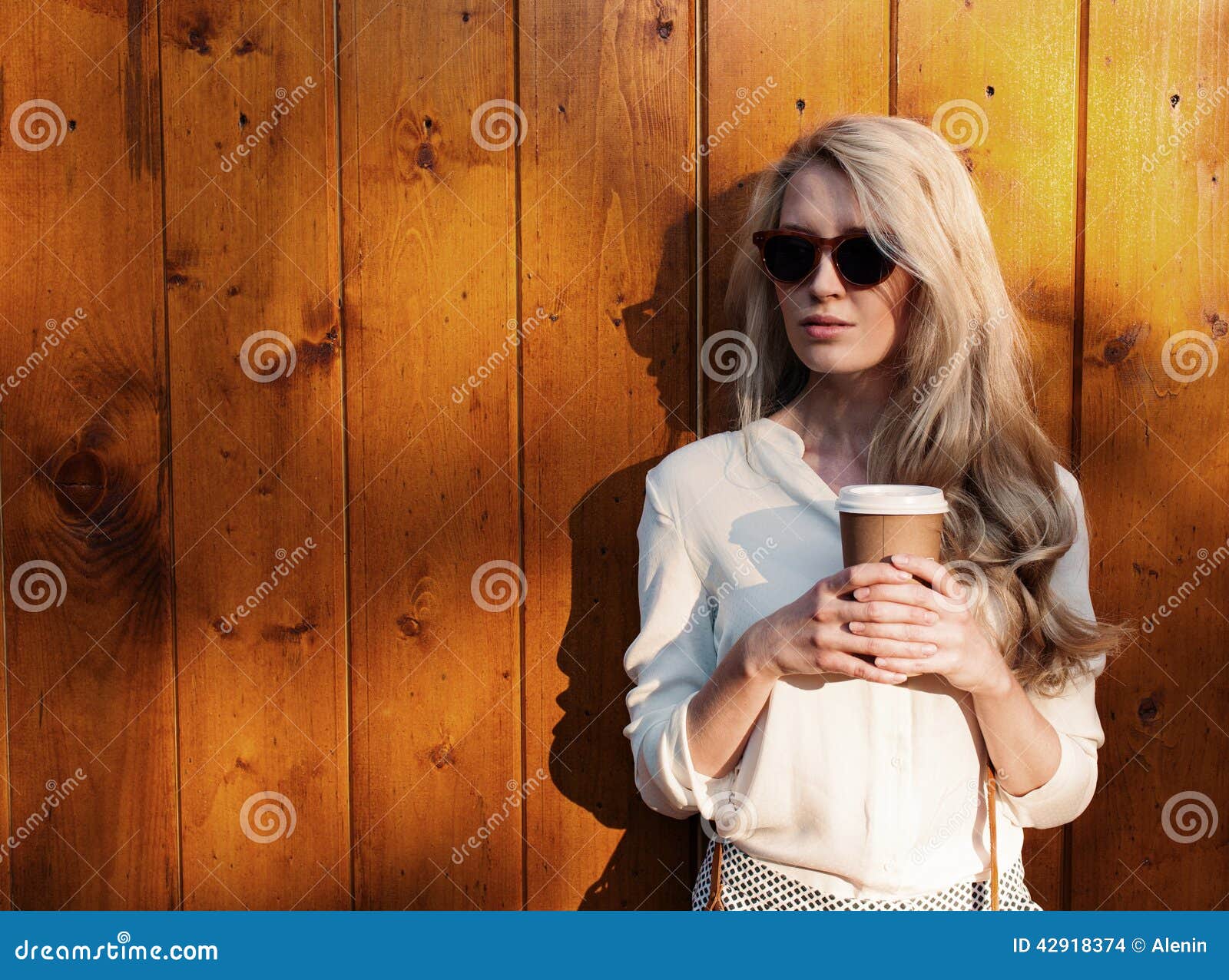 Junges sexy blondes Mädchen mit dem langen Haar in der Sonnenbrille, die einen Tasse Kaffee hält, haben den Spaß, der das weiche. Junges sexy blondes Mädchen mit dem langen Haar in der Sonnenbrille, die einen Tasse Kaffee glättet weiches Sonnenlicht hält