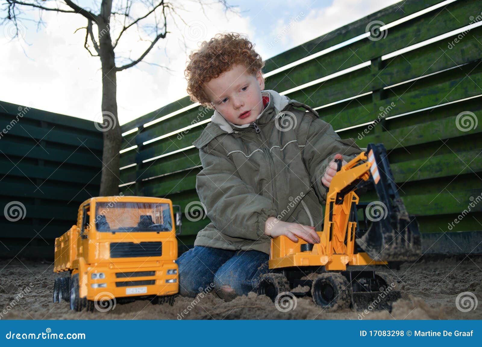 Kind mit Gebäudespielwaren. Junger Junge (5) in einem Garten, spielend mit einem Spielzeug-LKW und -exkavator im Sand