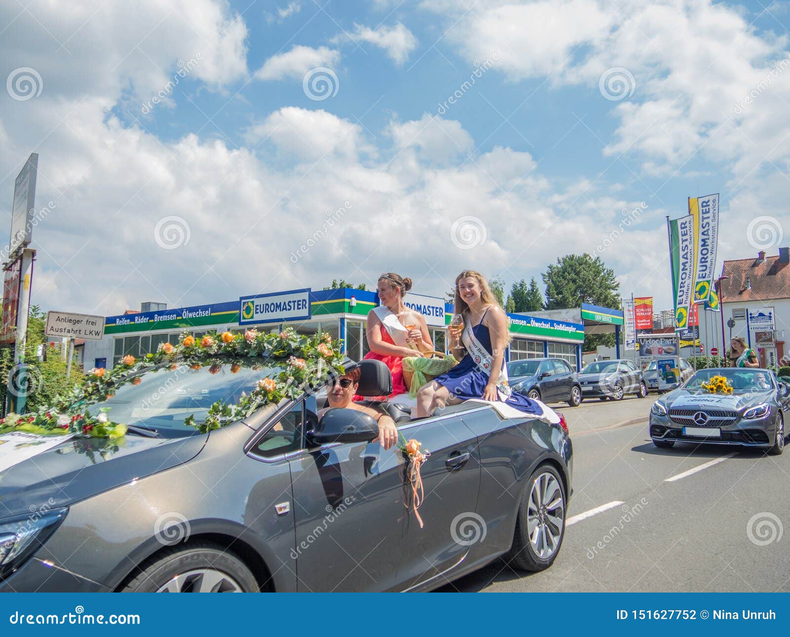 Kirchheimbolanden, Rheinland-Pfalz, Germany-06 23 2019: Feiertagsparade auf Straßen der deutschen Stadt während der Bier-Festival. Kirchheimbolanden, Rheinland-Pfalz, Germany-06 23 2019: Verzierter Cabriolet, der 2 lächelnde Mädchen in den Cocktailkleidern sitzen auf die Oberseite transportiert, wenn die Gläser Wein in den Händen, von einem anderen Auto gefolgt sind, der Parade