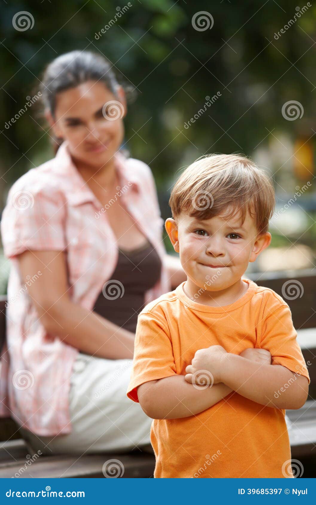 Kleiner Junge mit den stehenden Armen der Mutter gekreuzt. Kleiner glücklicher zufälliger kaukasischer Junge mit den stehenden Armen der Mutter gekreuzt. Elternteil im Hintergrund, lächelnd. Betrachten der Kamera.