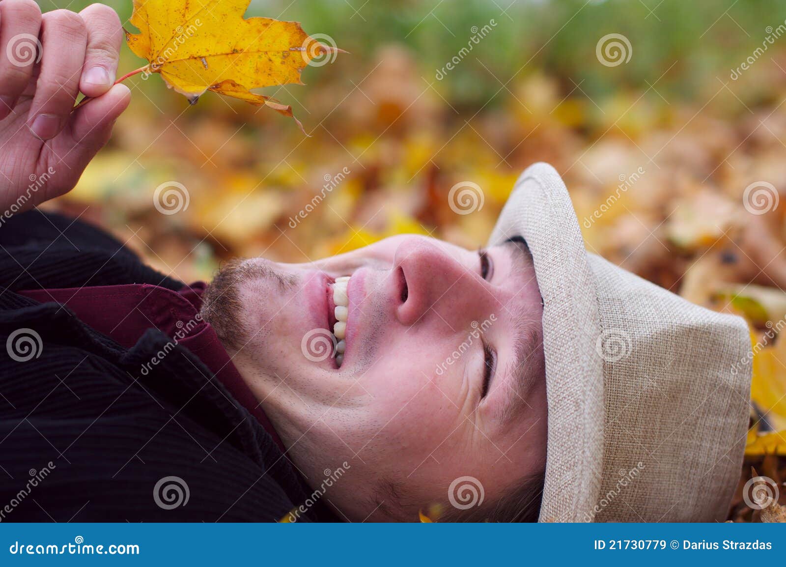 Knappe mens die en in de herfstbladeren glimlacht ligt. Knappe mens met hoed die in de herfstbladeren achtergrond en het glimlachen ligt. Houdend één verlof in hand