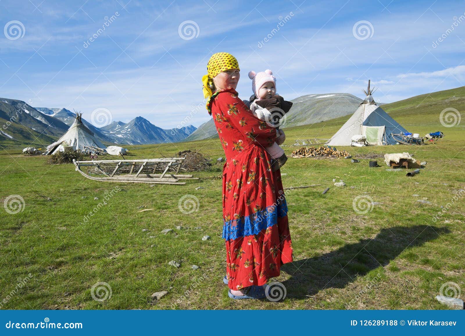 YAMAL RYSSLAND - AUGUSTI 27, 2018: Kvinnlign Hanty med barnet på händer mot bakgrunden av en bosättning av renavelsdjur
