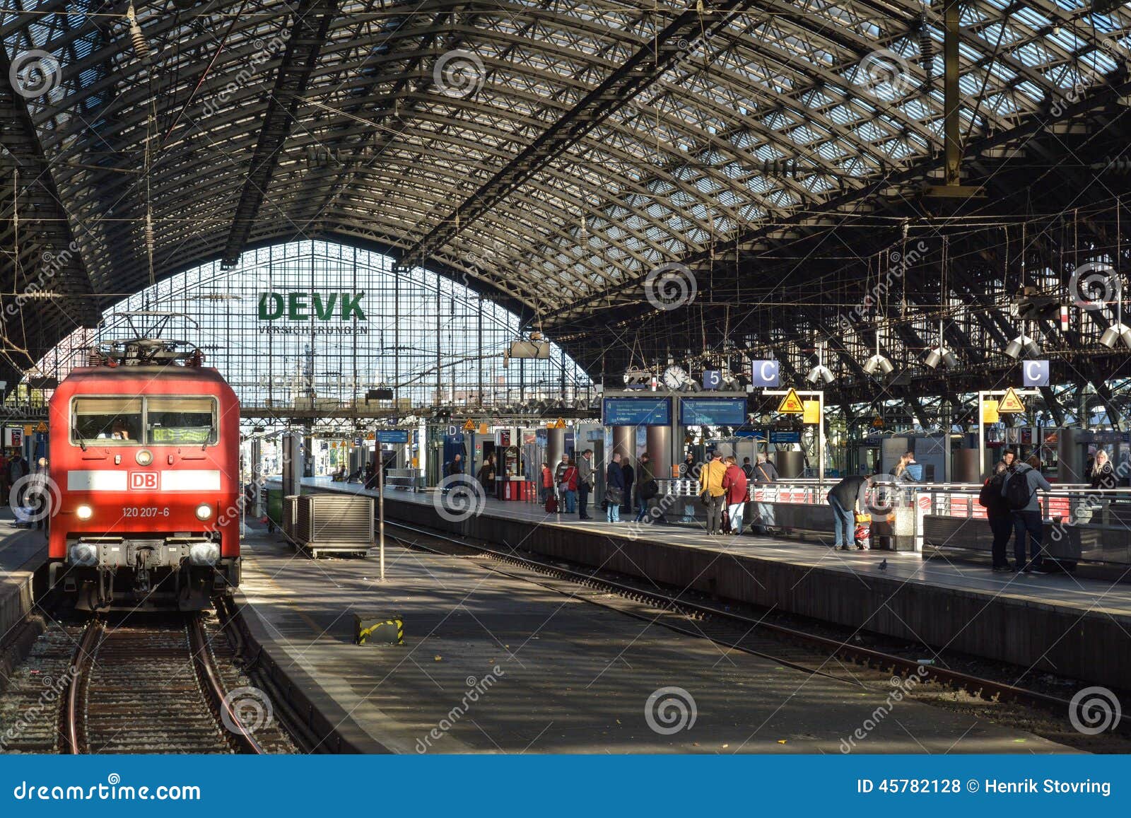 KÃ¶ln Hauptbahnhof (3), Colonia, Germania. Il corridoio principale della stazione ferroviaria principale in Colonia, Germania con un treno rosso dalla partenza aspettante di Deutsche Bahn - la luce solare brillante piove a dirotto tramite il tetto d'acciaio e di vetro sulle piste e sui binari