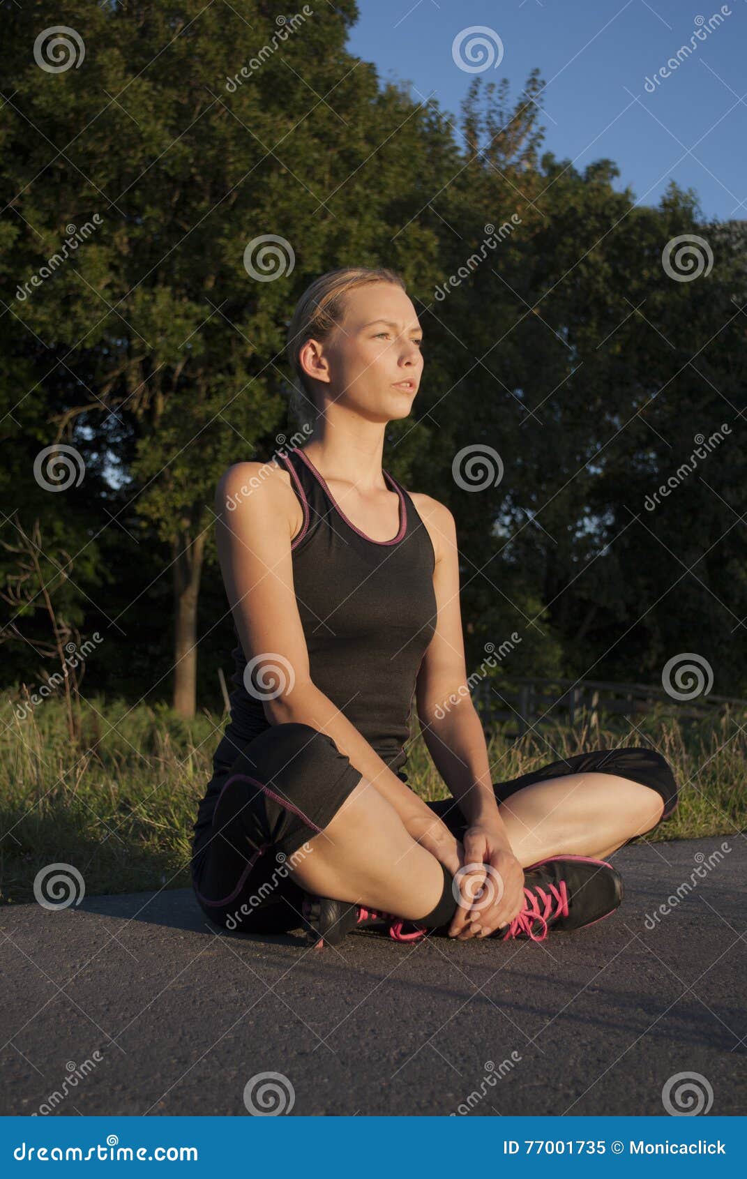 La belle jeune femme adulte sportive fait une pause pendant l'exercice extérieur, reposant à jambes croisé. La belle fille blonde caucasienne sportive, repose à jambes croisé à la route dans les vêtements de sport au coucher du soleil, prenant un moment pour refroidir et faisant des exercices de respiration