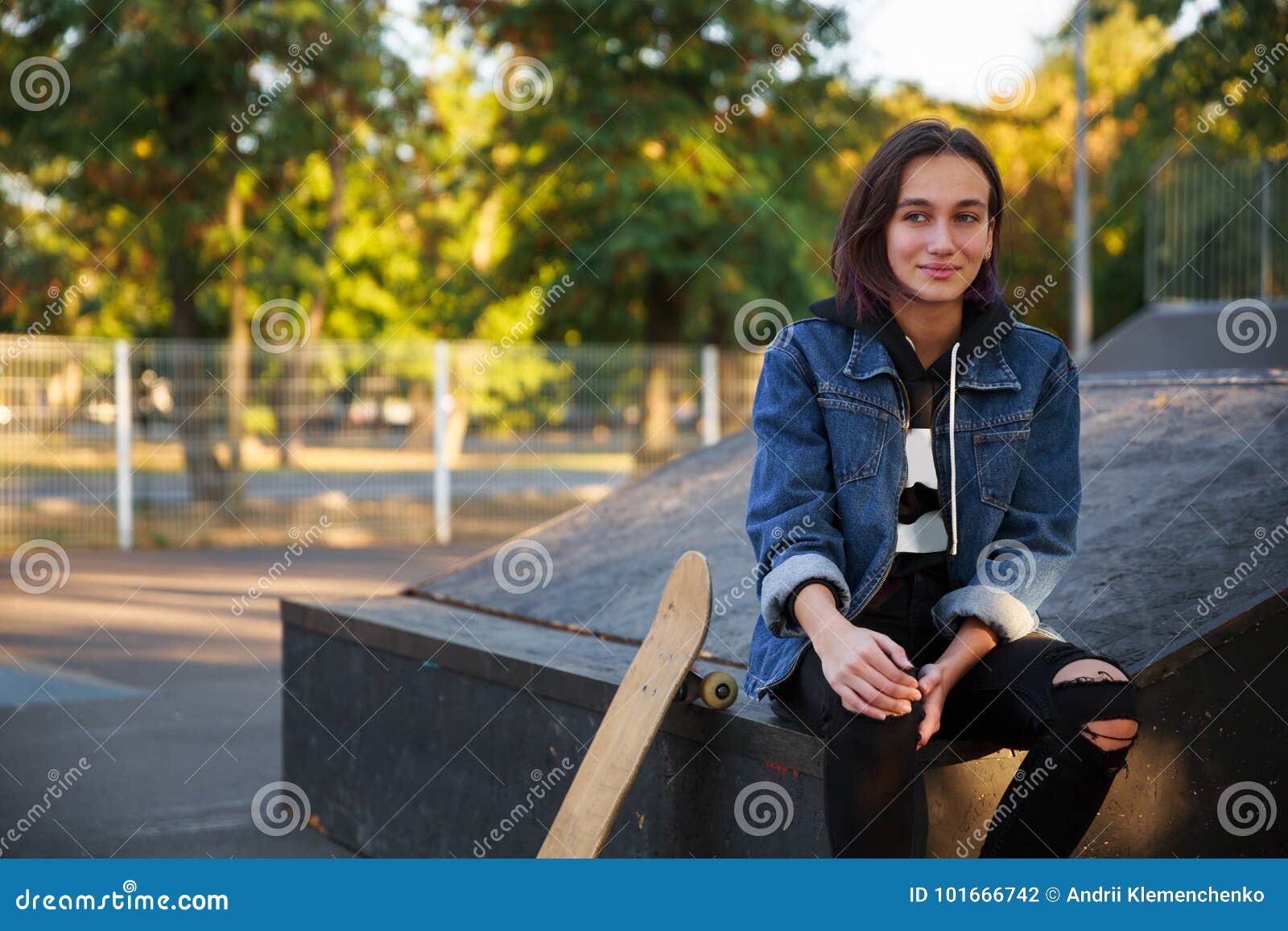 La chica joven hermosa se sienta al aire libre con un monopatín. Una chica joven hermosa en una chaqueta de los vaqueros se está sentando en el aire fresco con un monopatín Contra la perspectiva del parque Primer Concepto de ocio