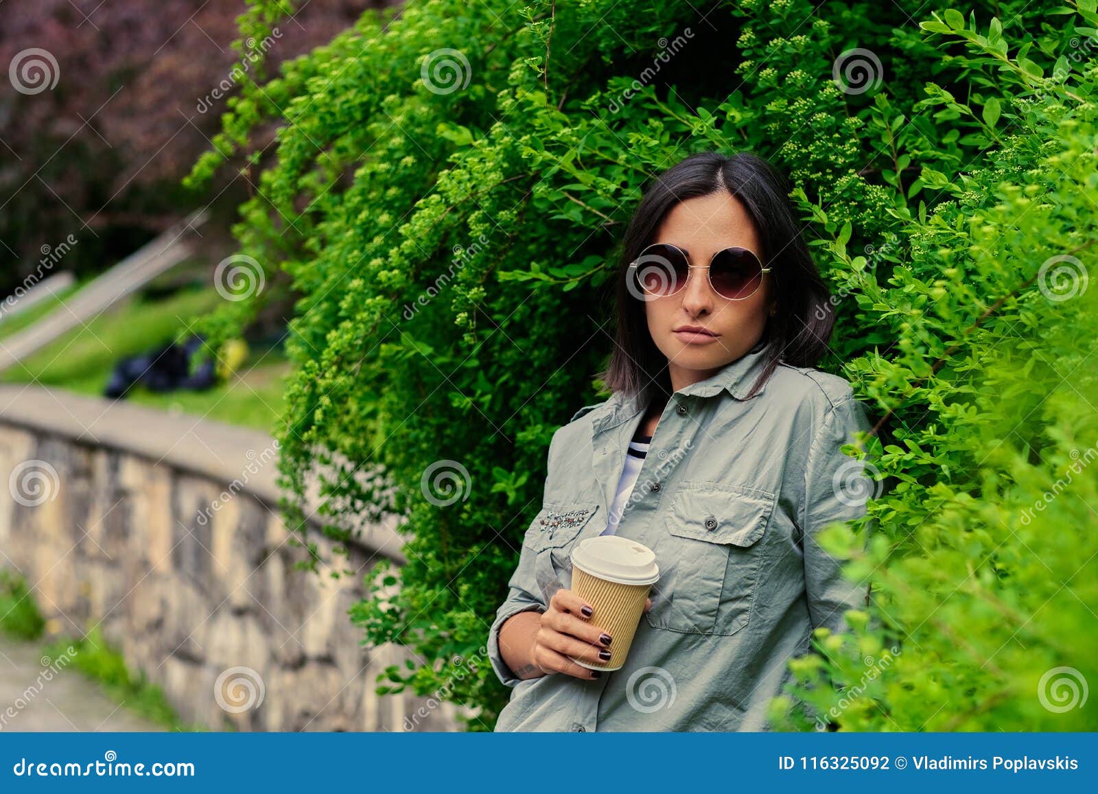 La femmina castana in occhiali da sole beve il caffè in un parco dell'estate