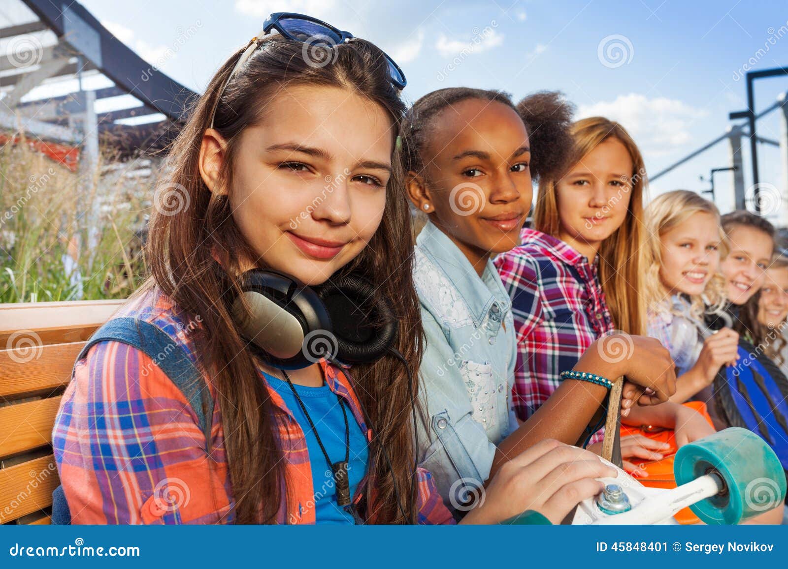 La muchacha con los auriculares y los amigos se sientan juntos. La muchacha hermosa con los auriculares y sus amigos se sientan en banco juntos en verano durante día con la construcción metálica en fondo y con el monopatín