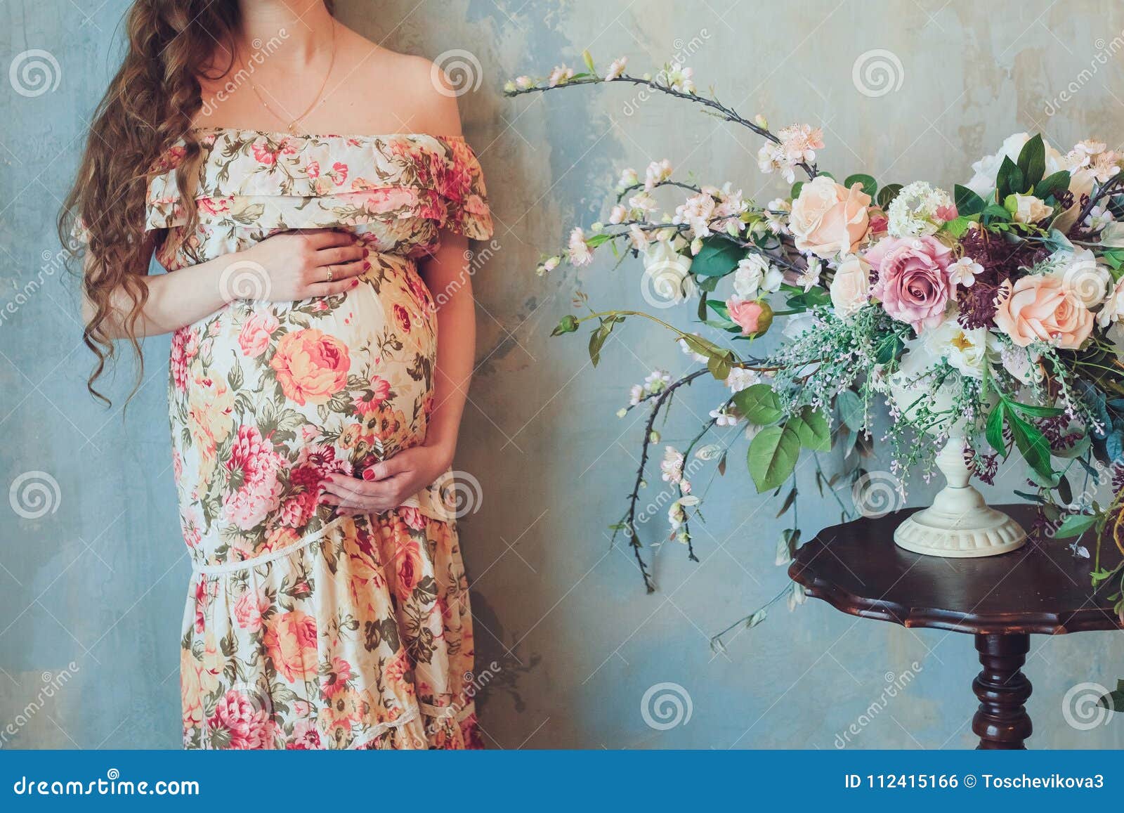 La Mujer Embarazada En Un Vestido Hermoso Se Está Colocando Lado De Un Ramo Brillante De Flores Y Lleva a Cabo Las Ma Foto de archivo - Imagen de sano,