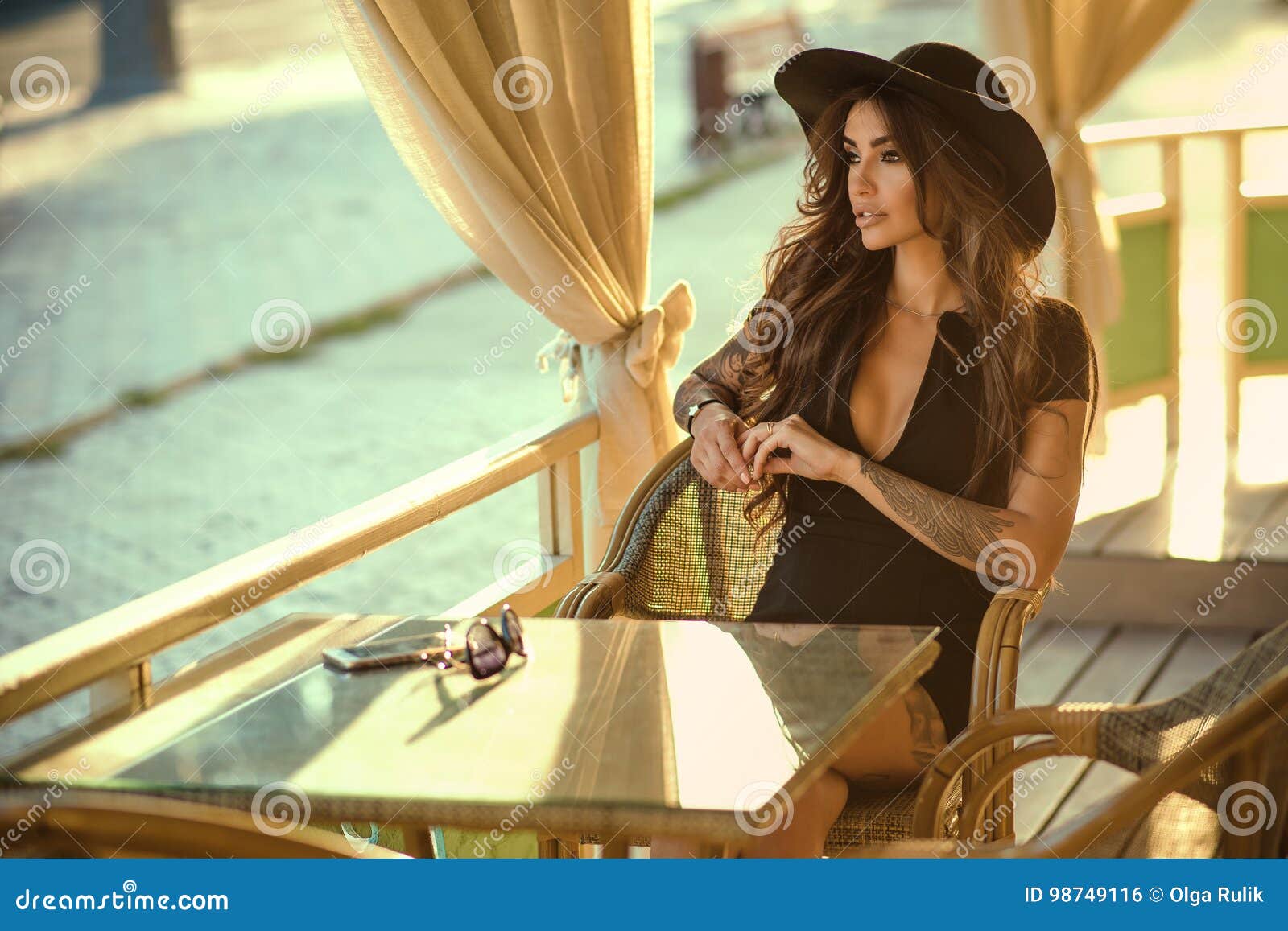 ÃÅ¸lam tattooed brunette in little black dress and trendy wide brimmed fedora hat sitting in the nice summer restaurant