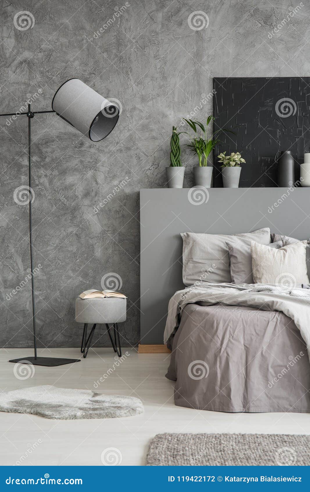 lamp and stool next to bed with pillows and bedhead in grey bedroom interior with poster. real photo