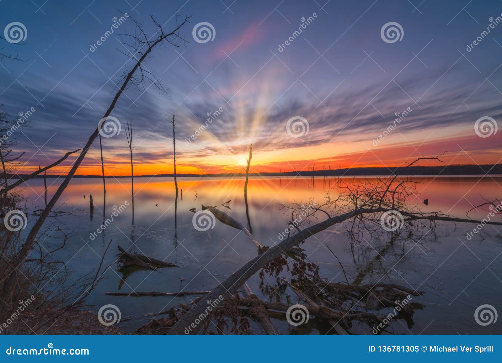 Langer Belichtungssonnenaufgang mit Wolkenbewegung an Manasquan-Reservoir. Die schöne Ansicht des Eises bildend entlang dem Wasser umranden nahe gefallenen Klotz mit während einem bunten Sonnenaufgang