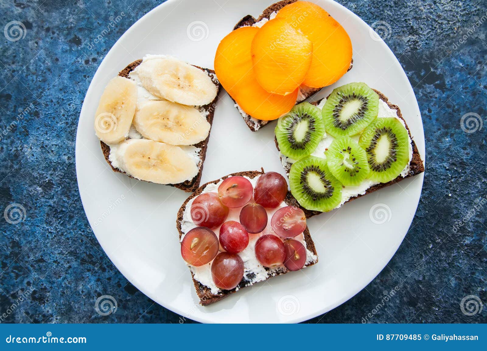 Las rebanadas de la variedad de pan de centeno tuestan con las frutas Caqui del plátano,. Las rebanadas de la variedad de pan de centeno tuestan con las frutas Bocadillos de la uva del kiwi del perssimon del plátano en una placa blanca en fondo oxidado