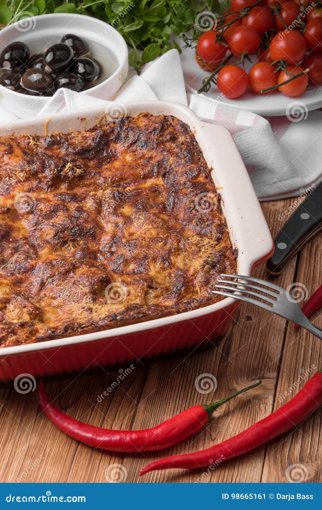 Lasaña cocida fresca en plato rojo con los tomates y los chiles de las aceitunas negras en la tabla de madera con las hierbas y la bifurcación