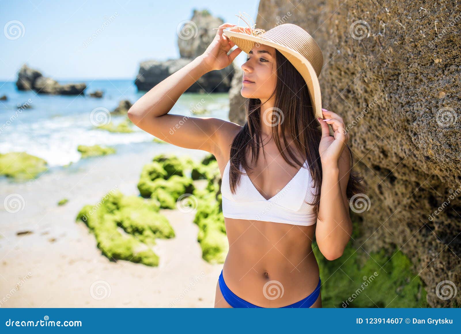 Latijns mooi gelukkig meisje in bikinibovenkant en strohoed die zich met gesloten ogen op rotsachtig strand bevinden. Hoge hoekmening van mooi gelukkig meisje in bikinibovenkant en strohoed die zich met gesloten ogen op rotsachtig strand bevinden