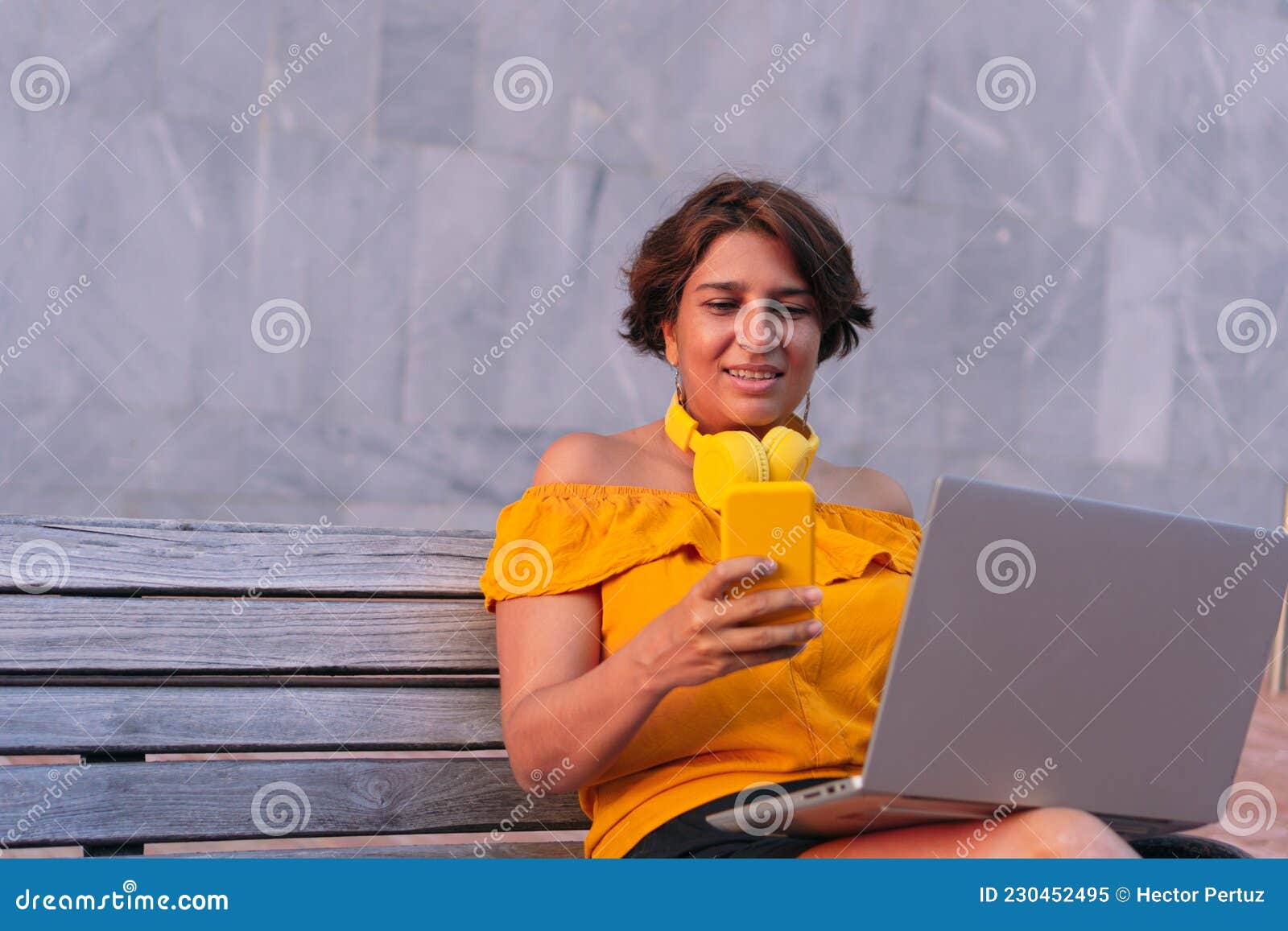 Latin Mature Woman Using A Cell Phone And Laptop While Sitting On A Bench In The Park Stock