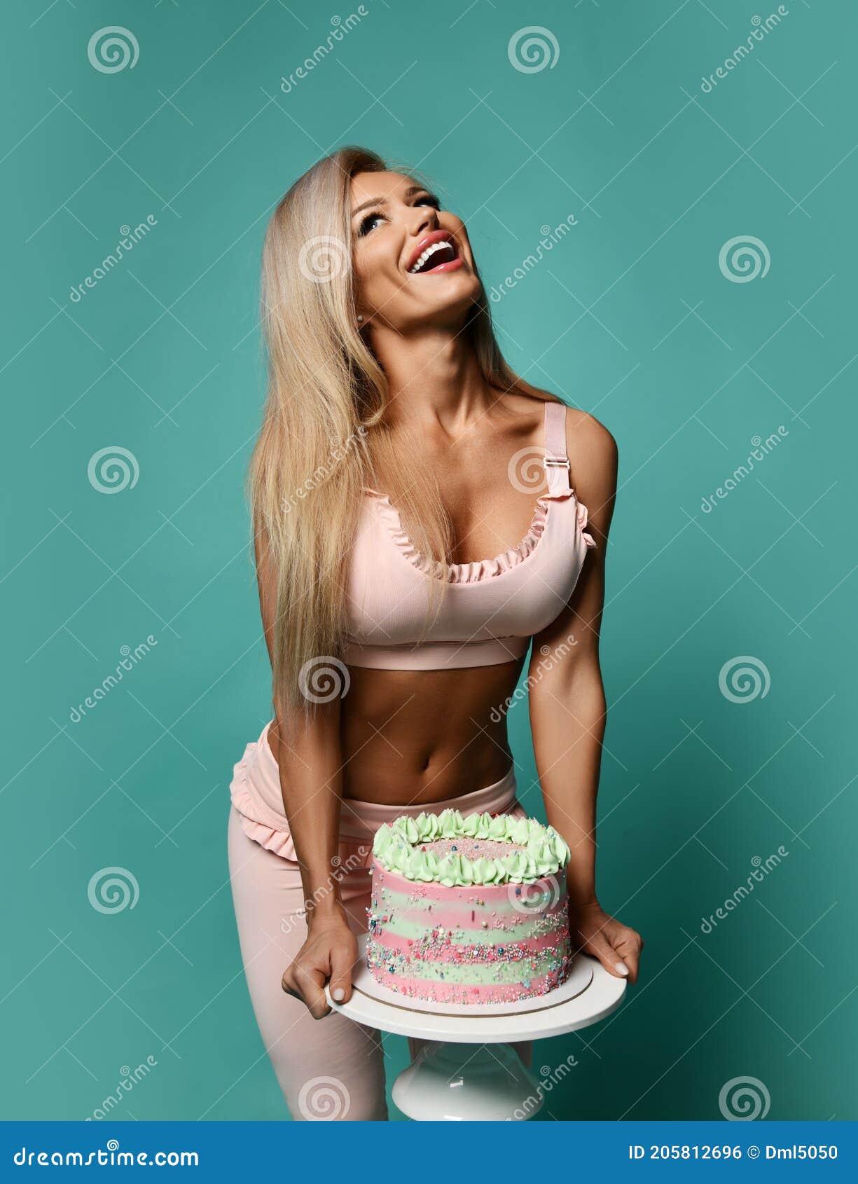 Laughing Blonde Fitness Woman in Top Bra and Pants Stands Holding Birthday Holiday Cake in Hands and Looks Up Stock Photo picture