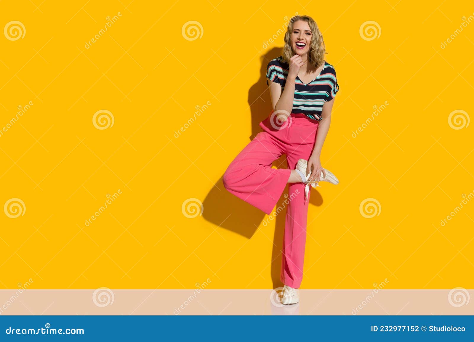 laughing young woman in pink high waisted baggy pants and sneakers is posing on one leg in front of yellow wall