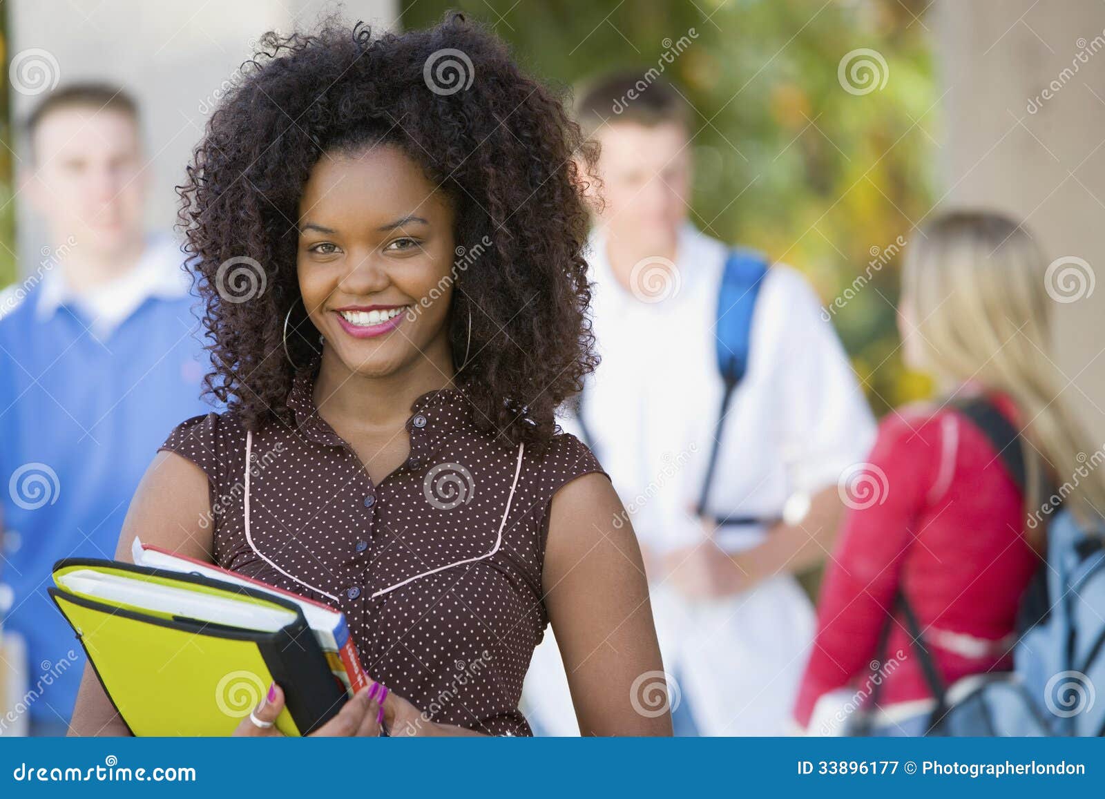 Le den kvinnliga studenten On College Campus. Stående av att le den kvinnliga studenten för afrikansk amerikan på högskolauniversitetsområde med klasskompisar i bakgrund