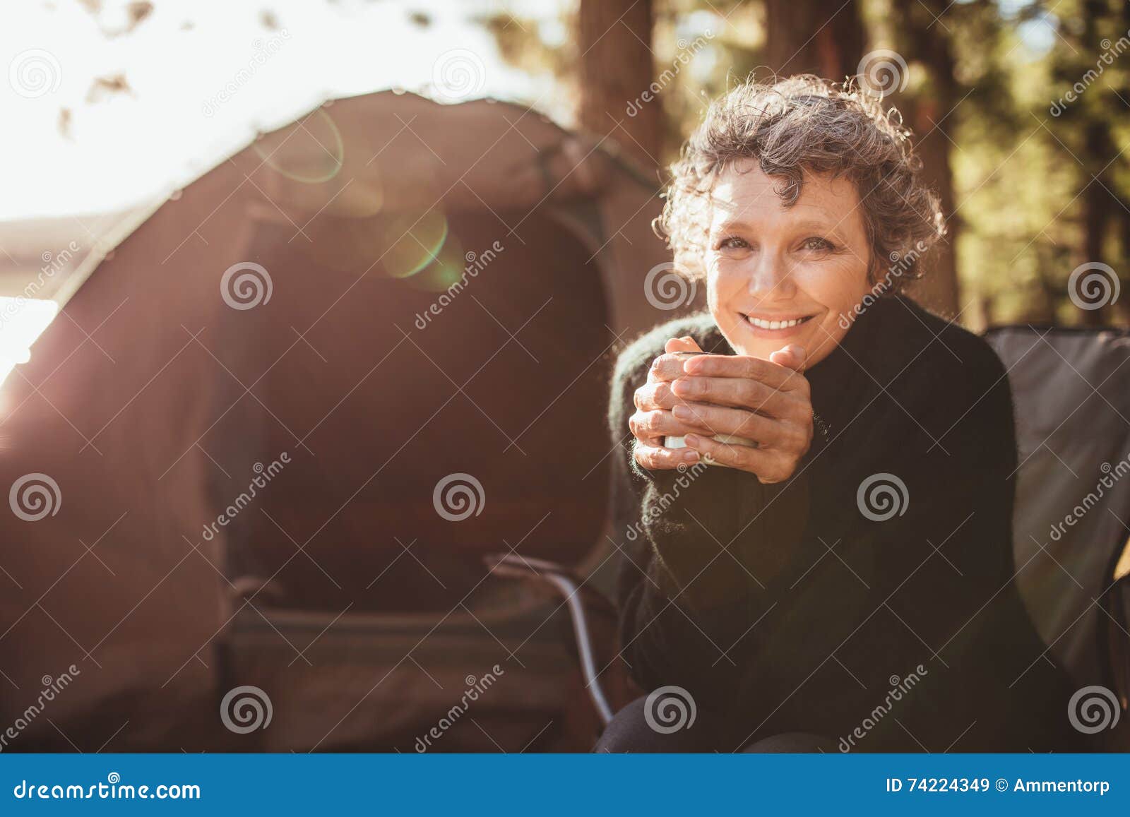 Le mogna par på att campa. Stående av en hållande kaffekopp för lycklig mogen kvinna, medan sitta framme av ett tält och se kameran Hög caucasian kvinna som har te på campingplatsen på en solig dag