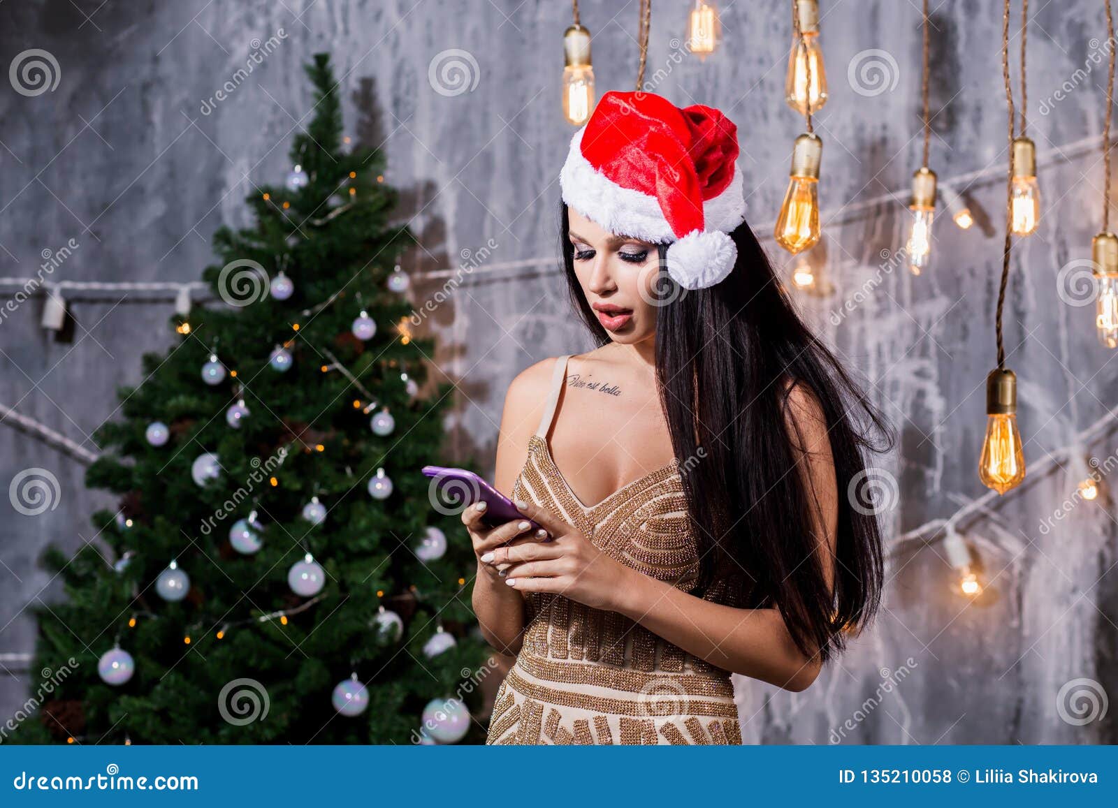 Le téléphone élégant de participation de femme regardant l'écran l'arbre de Noël s'allume sourire de port de chapeau de Santa Concept saisonnier de salutations L'espace pour le texte Vacances, nouvelle année, célébration