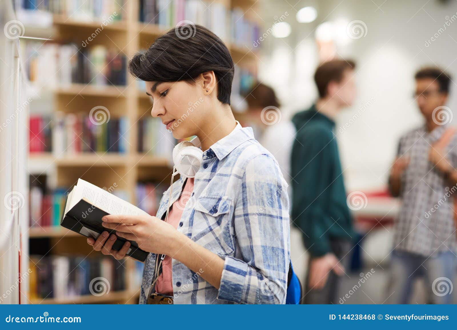 Lecture du contenu du livre. Étudiant universitaire féminin attirant réfléchi sérieux avec les écouteurs de câble sur la position de cou dans la teneur en librairie et en lecture du livre