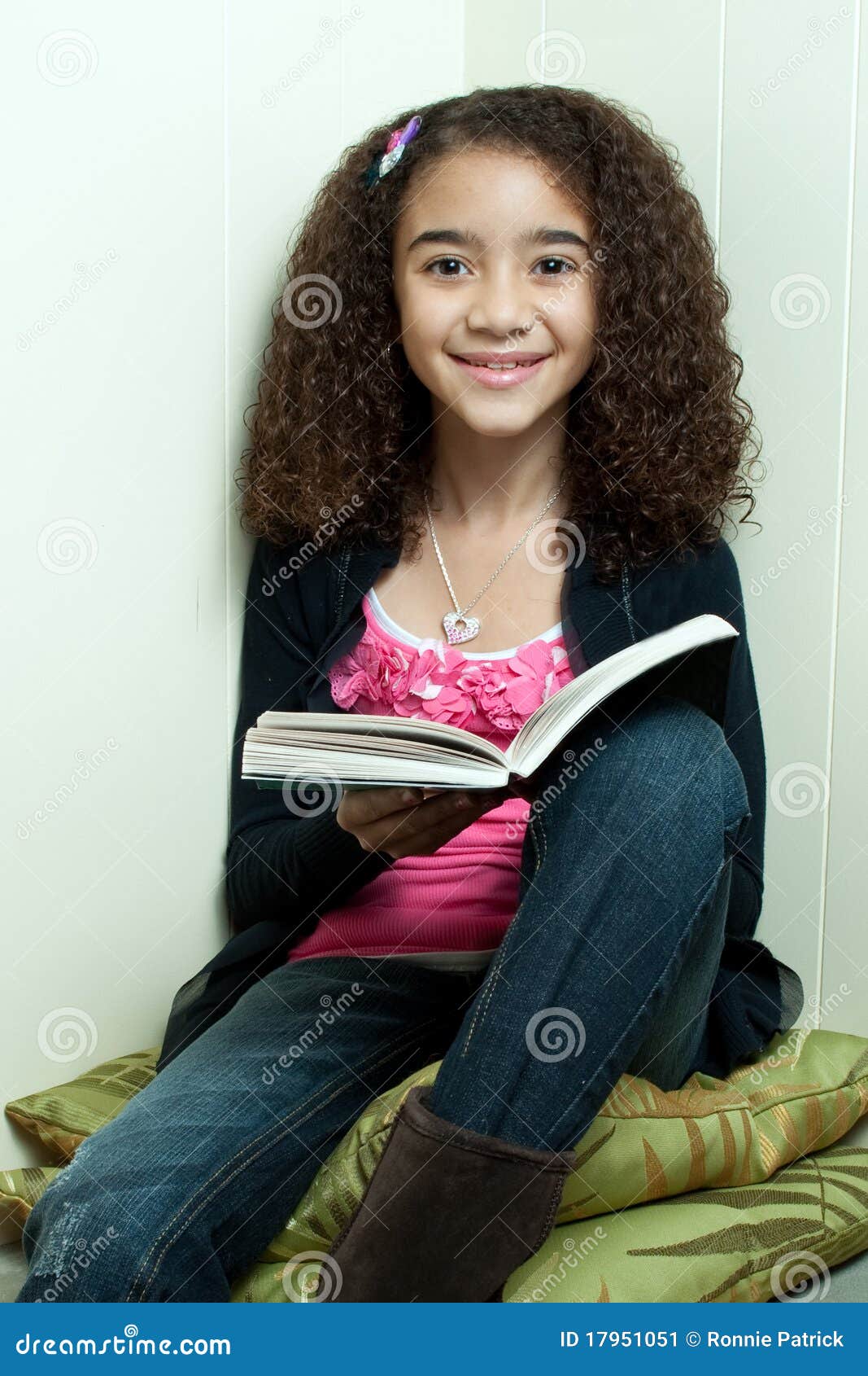 Leitura da rapariga no canto. Menina descasc escura nova com o cabelo curly, sentando-se em um canto em coxins, lendo um livro, sorrindo e olhando a câmera.
