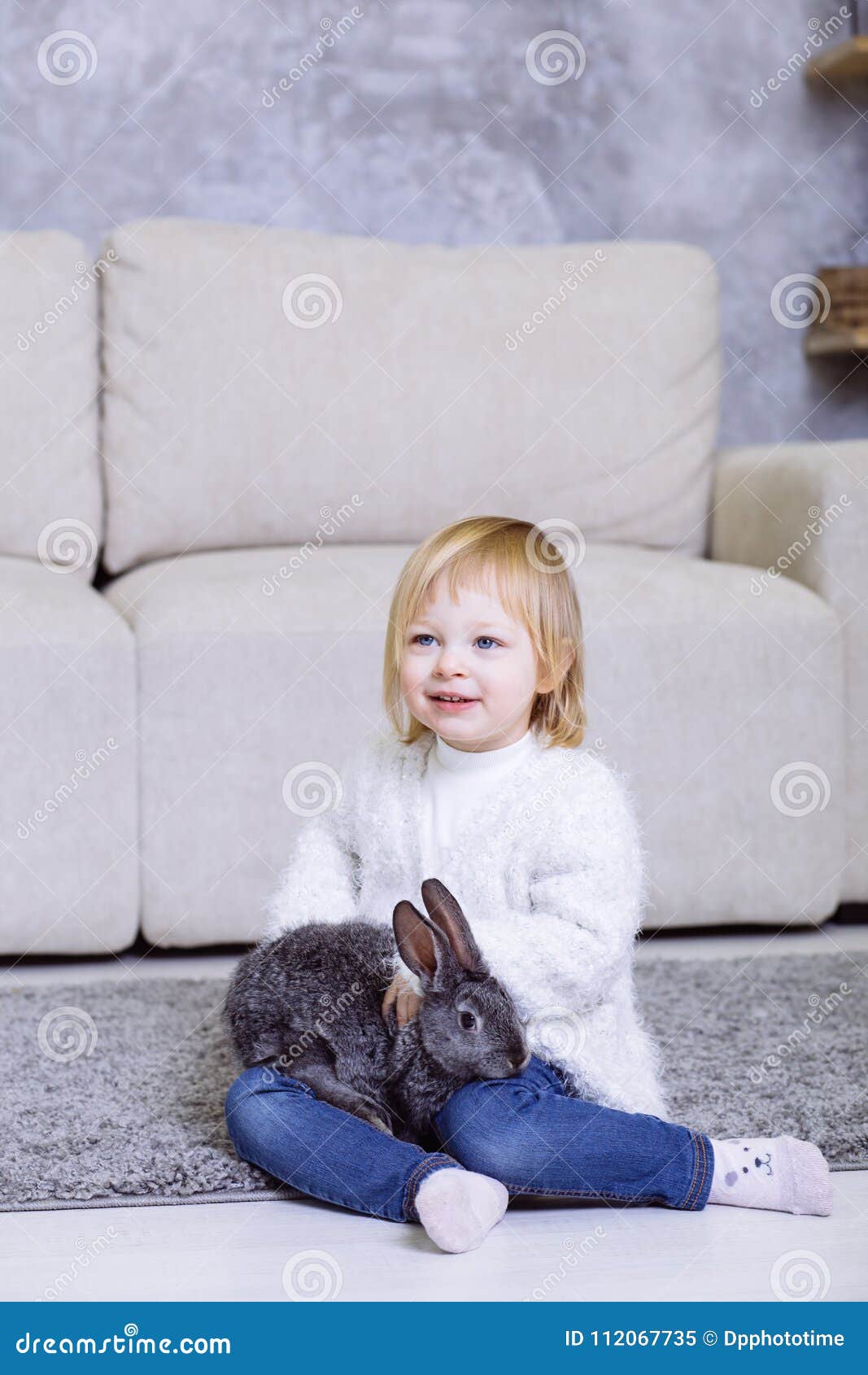 Leuk meisje met grijs Pasen-konijn, konijntje Het meisje van de blondebaby in witte sweater glimlachen en jeans die thuis op c zi. Leuk meisje met grijs Pasen-konijn, konijntje Het meisje van de blondebaby in witte sweater glimlachen en jeans die thuis op het tapijt dichtbij de bank zitten