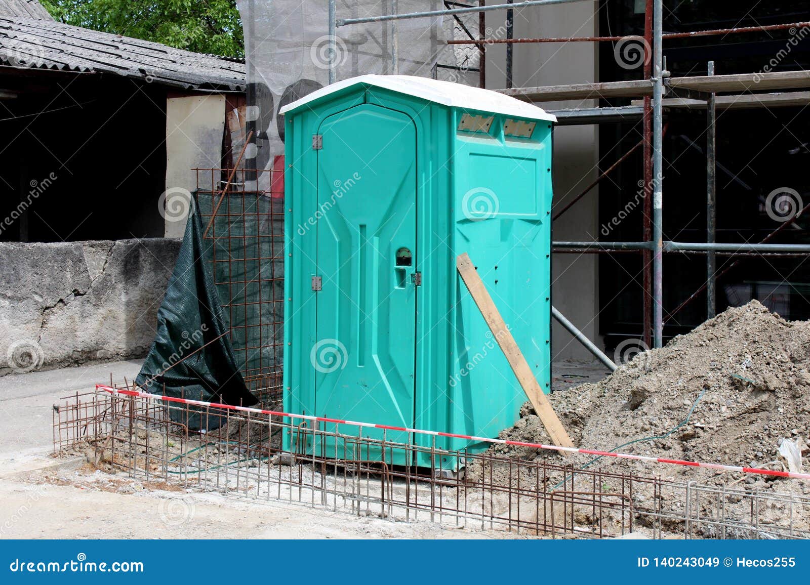 light green portable ecological toilet left on construction site surrounded with sand and other construction material with old