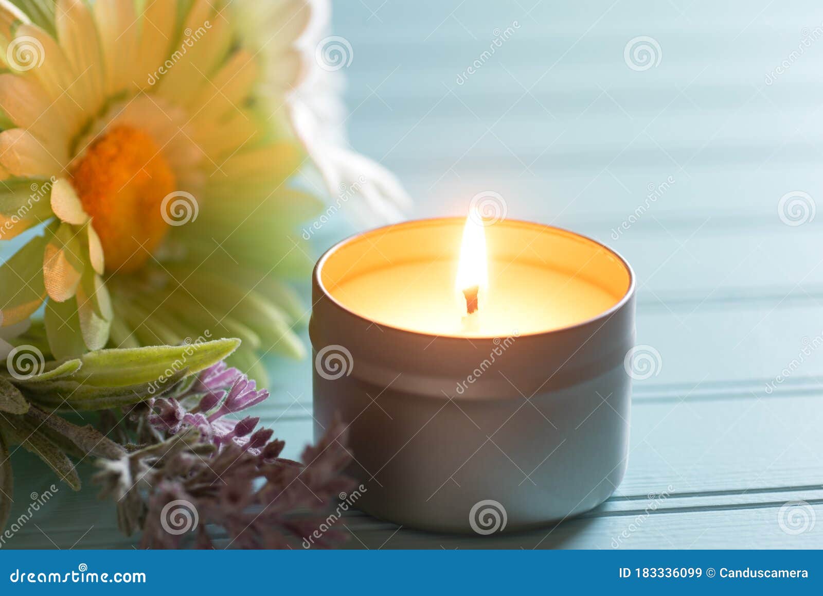 lighted and scented votive candle in a tin holder with flowers on the side, all on teal boards table background with window light