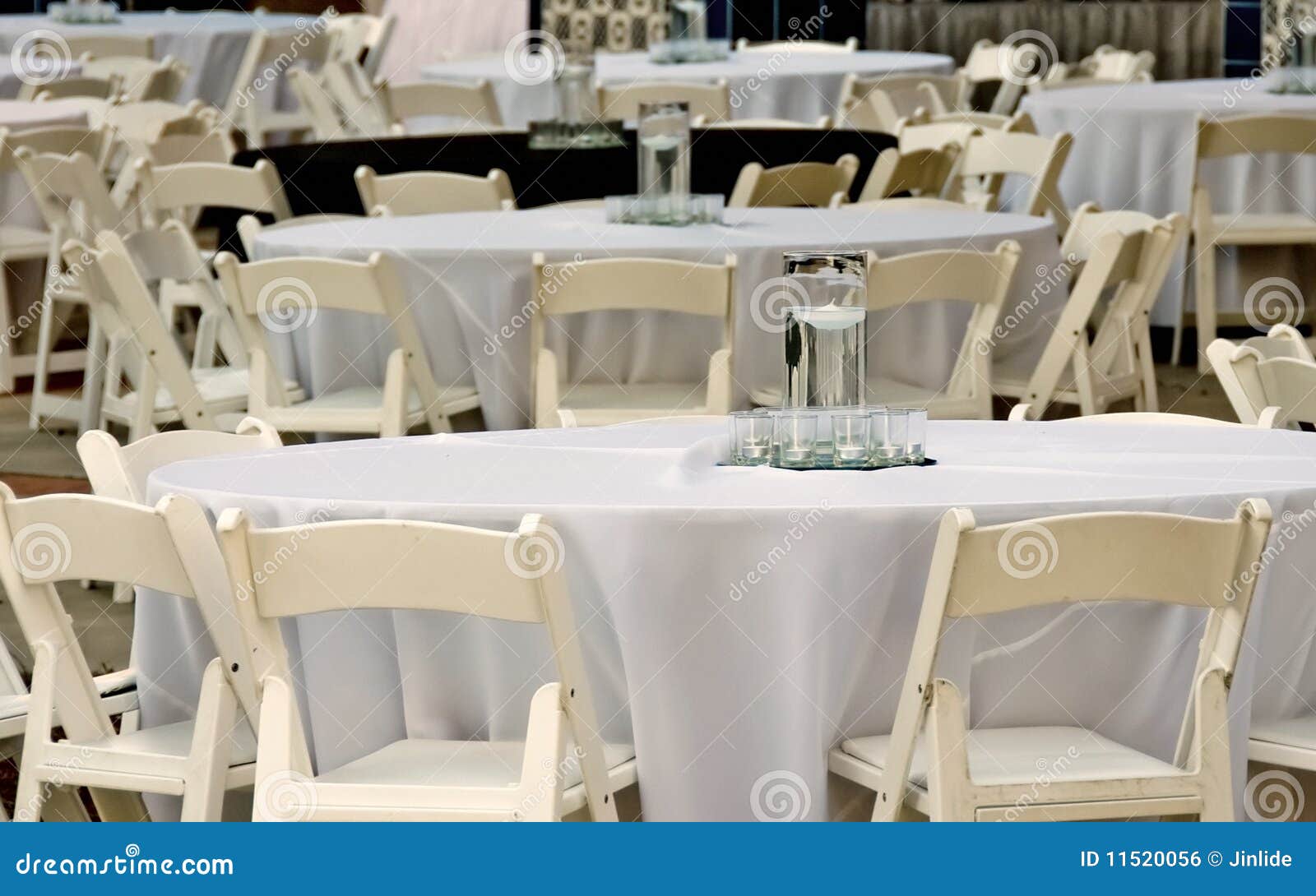 Lijsten en stoelen voor avondgebeurtenis. Vele bijeenkomsten met witte lijstkleren worden omringd door room-kleur houten vouwende stoelen. Glazen met water en drijvende kaarsen in het centrum van elke lijst.