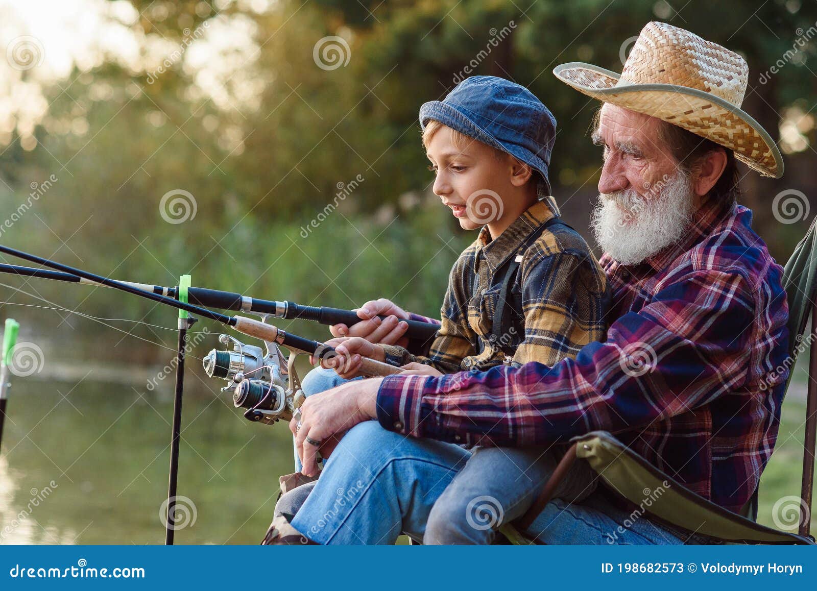 likable respected 70-aged bearded grandfather holding on knees 10-aged cute grandson and teaching him to catch fish.