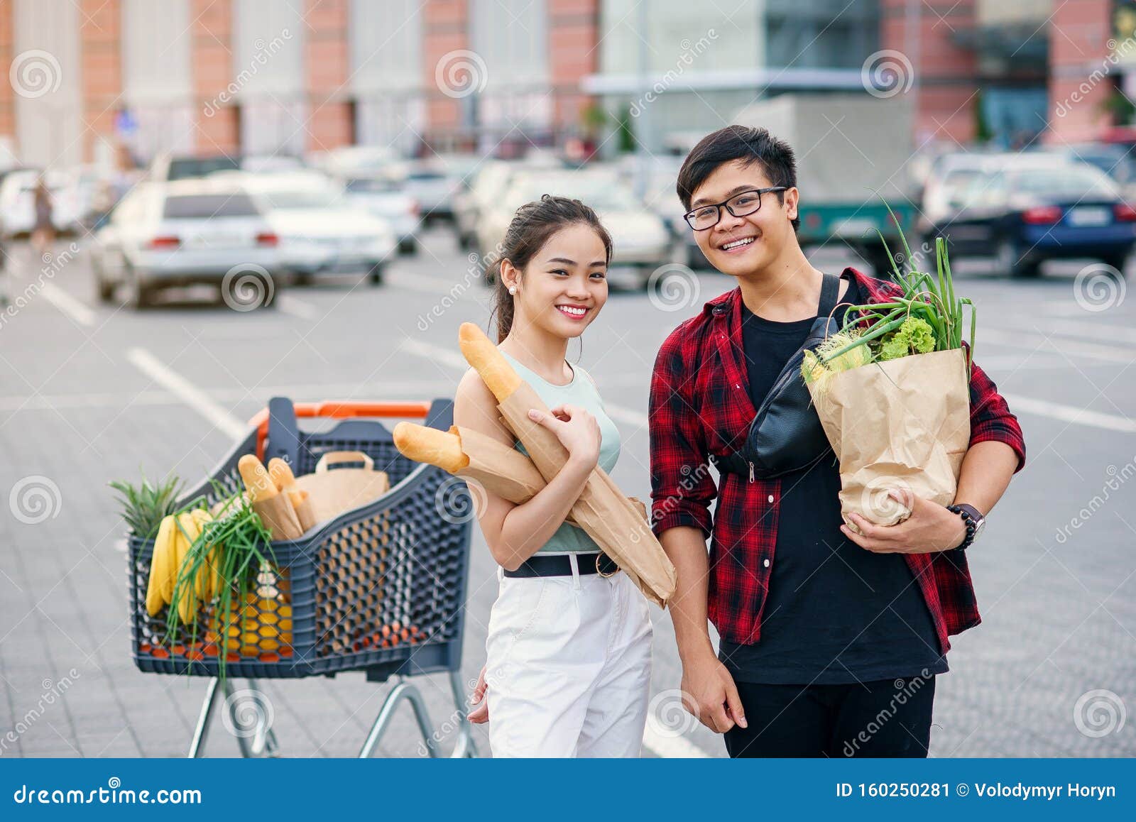 likable asian couple hold paper eco bags with organic healthy food in hands while standing near store mall. happy family