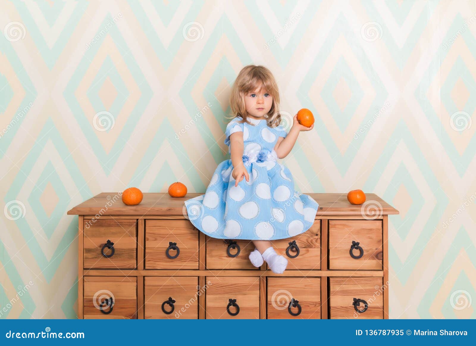Little Baby Girl Sitting Cross Legged On Chest Of Drawers Holding