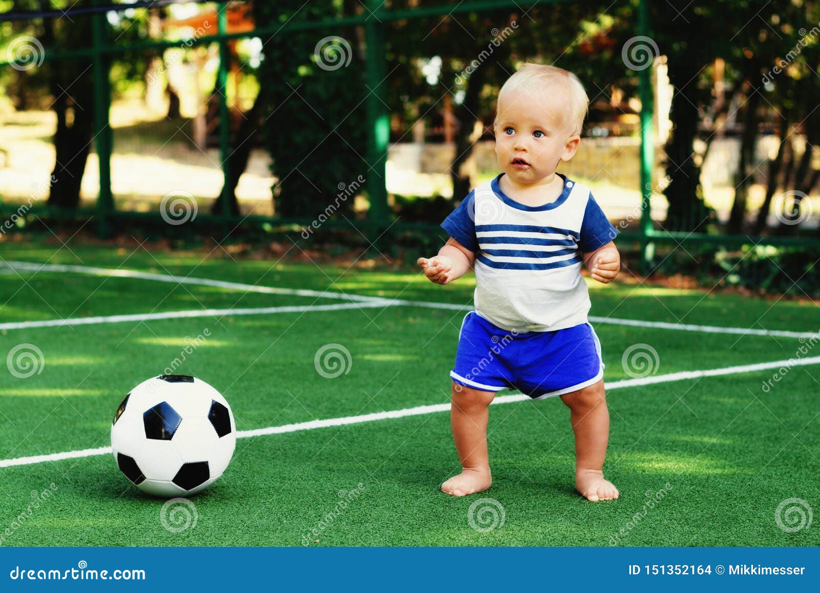 Little boy in blue shorts playing with soccer ball at sports ground. Blonde child in uniform standing at football field with ball. Little boy in blue shorts playing with soccer ball at sports ground. Blonde child in sports uniform standing at football field with a ball. Summer kids activity concept