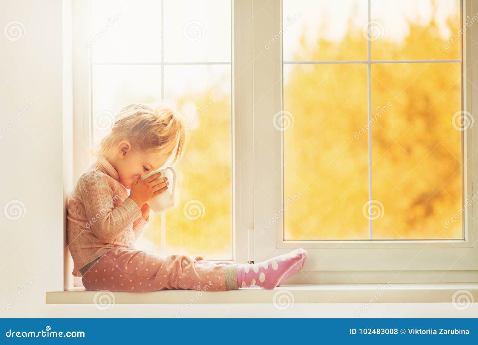 little cute kid girl sitting by window indoor holding cup of hot drink cocoa enjoying autumn forest background. season beauty