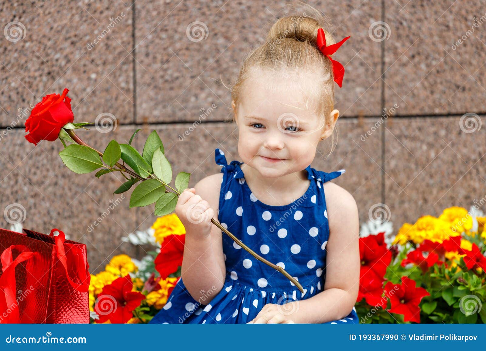 A Little Girl in a Blue Dress and a Red Bow Sits with Colored Bags on ...