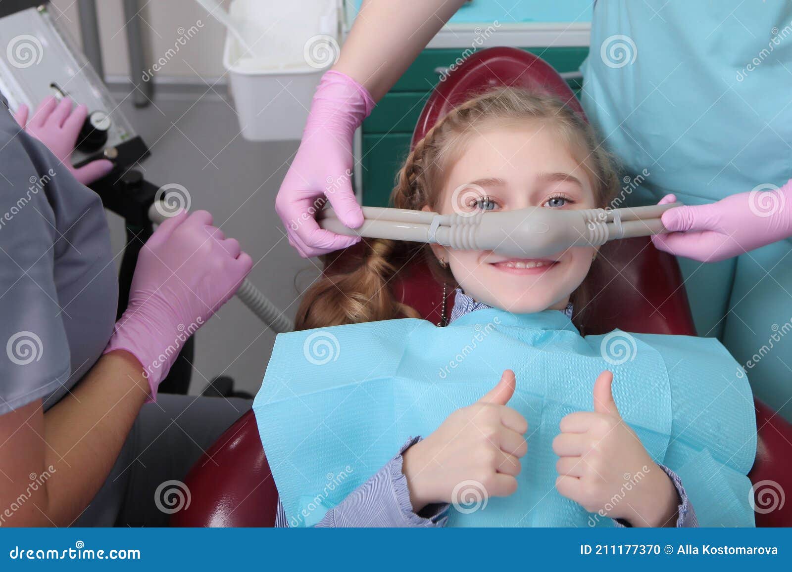 a little girl is comfortable to treat her teeth under superficial sedation. the girl smiles and holds two thumbs up. milk teeth