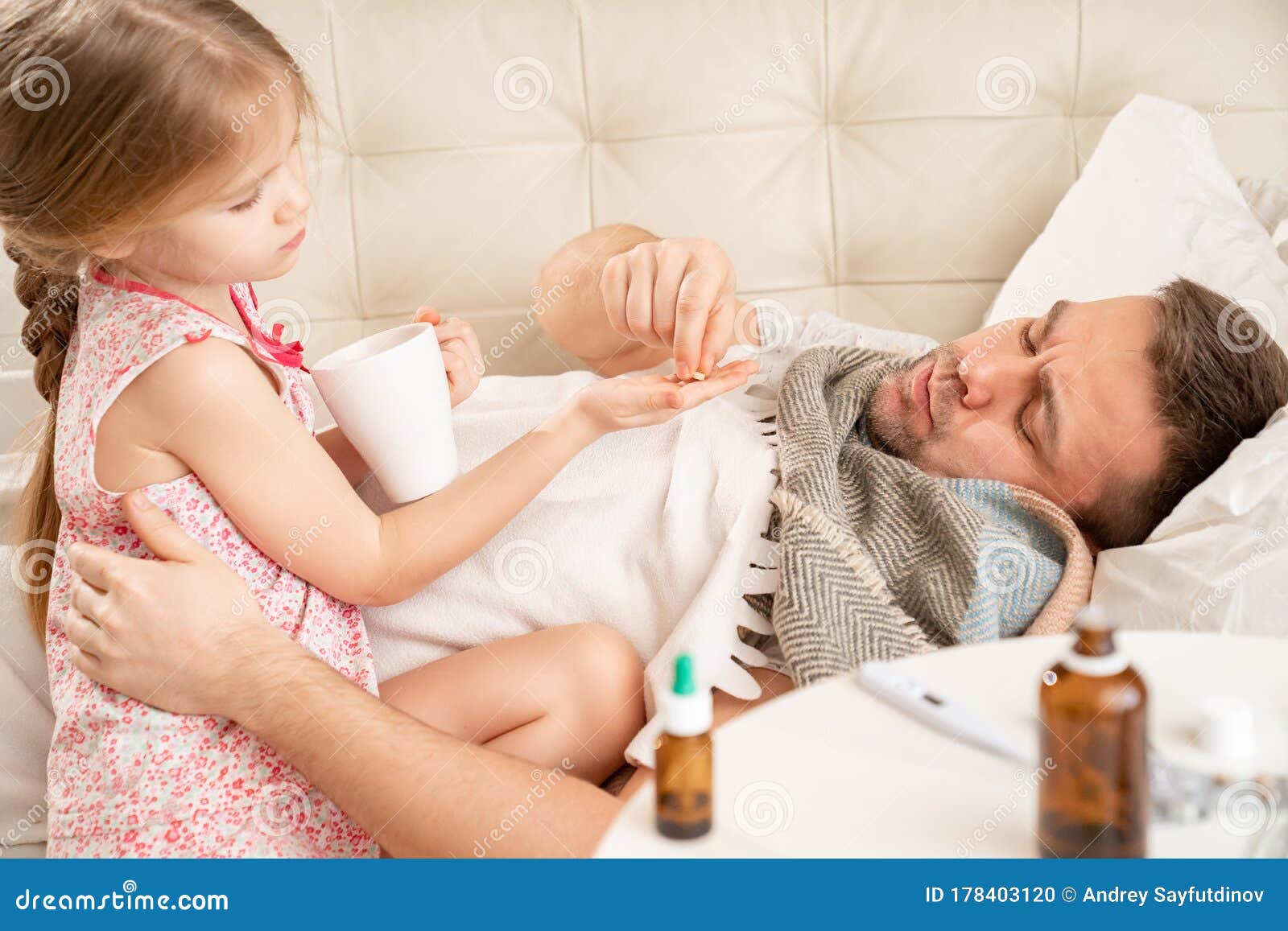  A little girl sits on the lap of her sick father who is lying in bed. She is holding a cup and a pill in her hands.