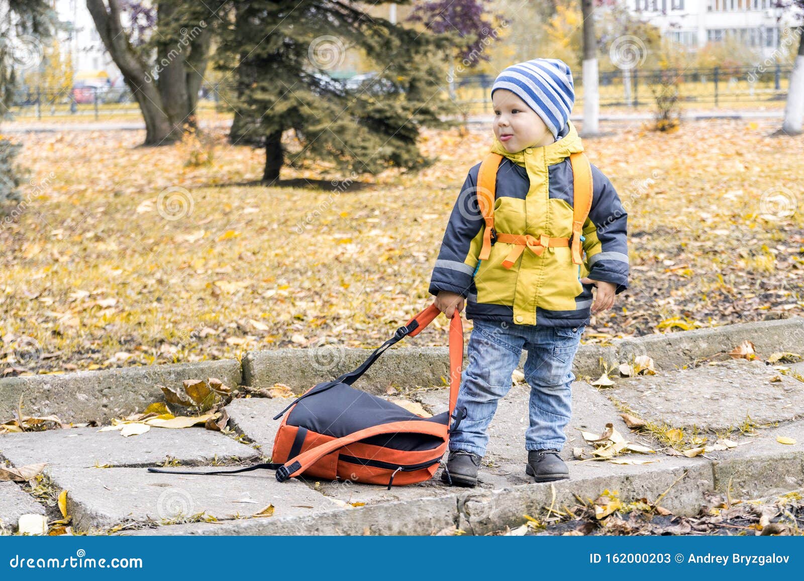 little hooligan boy dragged away his mother`s backpack and make grimace at her in autumn city park
