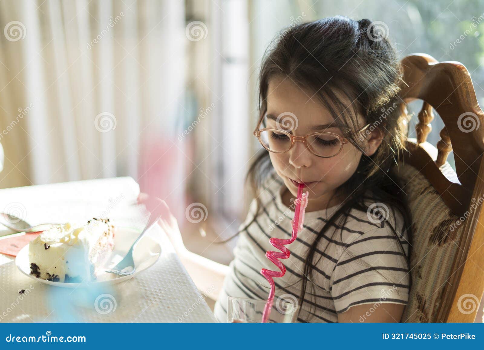 a little sad girl with glasses sits at the table in front of a plate with cake and drinks a drink through a straw.