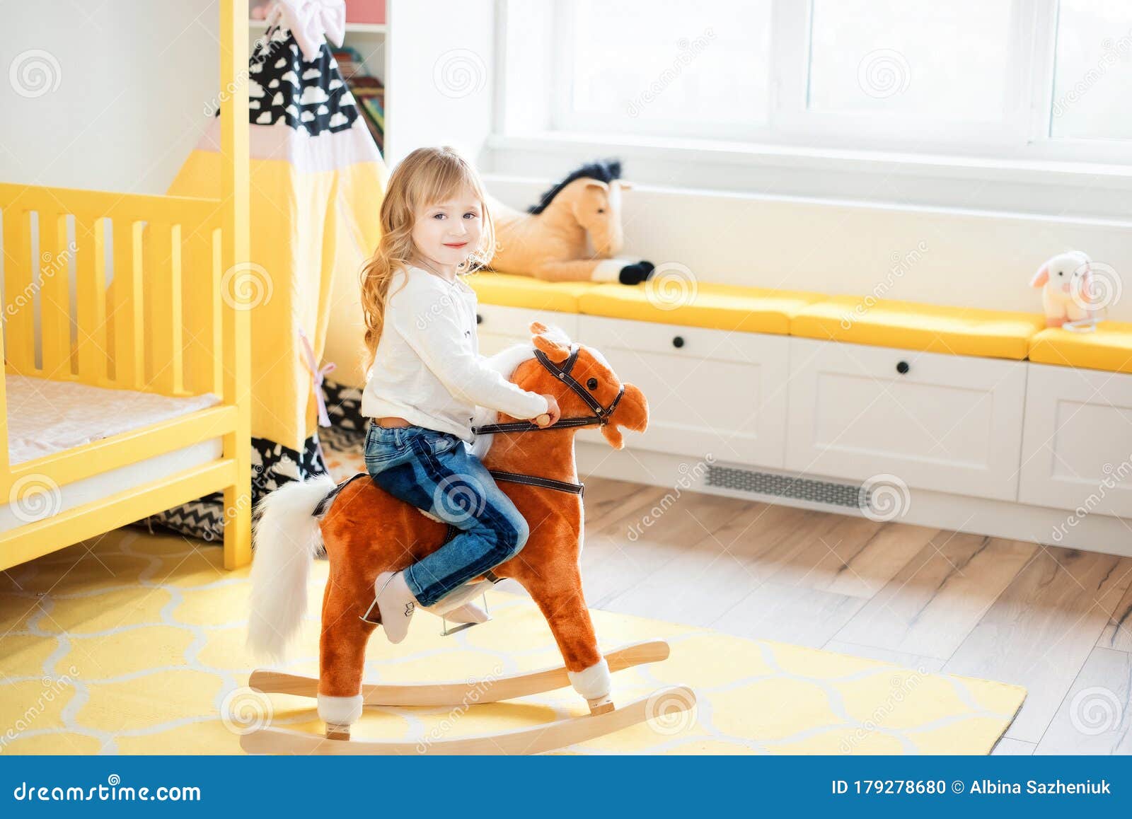 little smiling blonde 3 years old girl sitting on rocking horse in yellow bedroom at home in day time. time to play during self