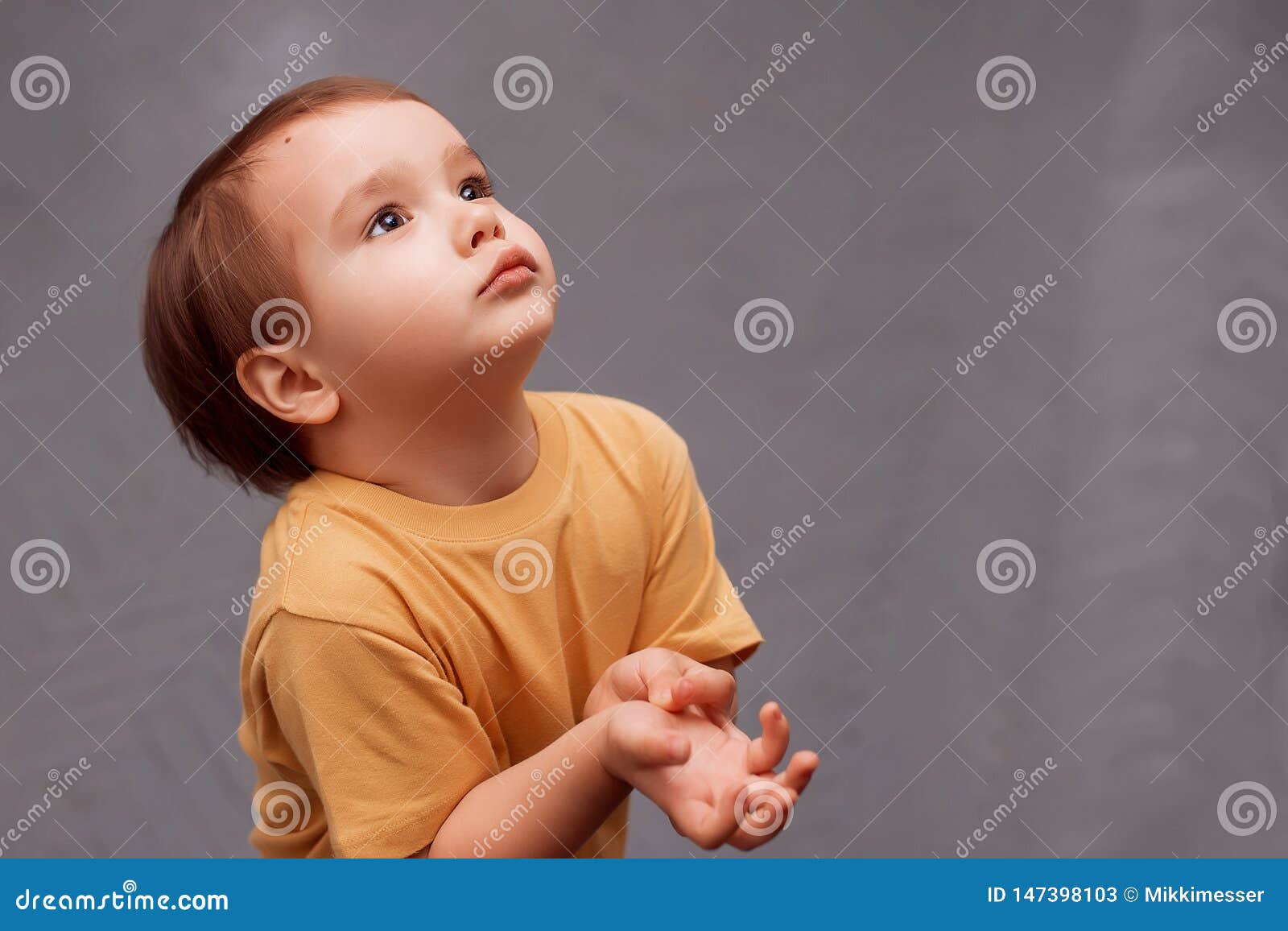 little toddler boy in yellow shirt standing and looking up asking for something. the kid is holding hands raised to chest like he