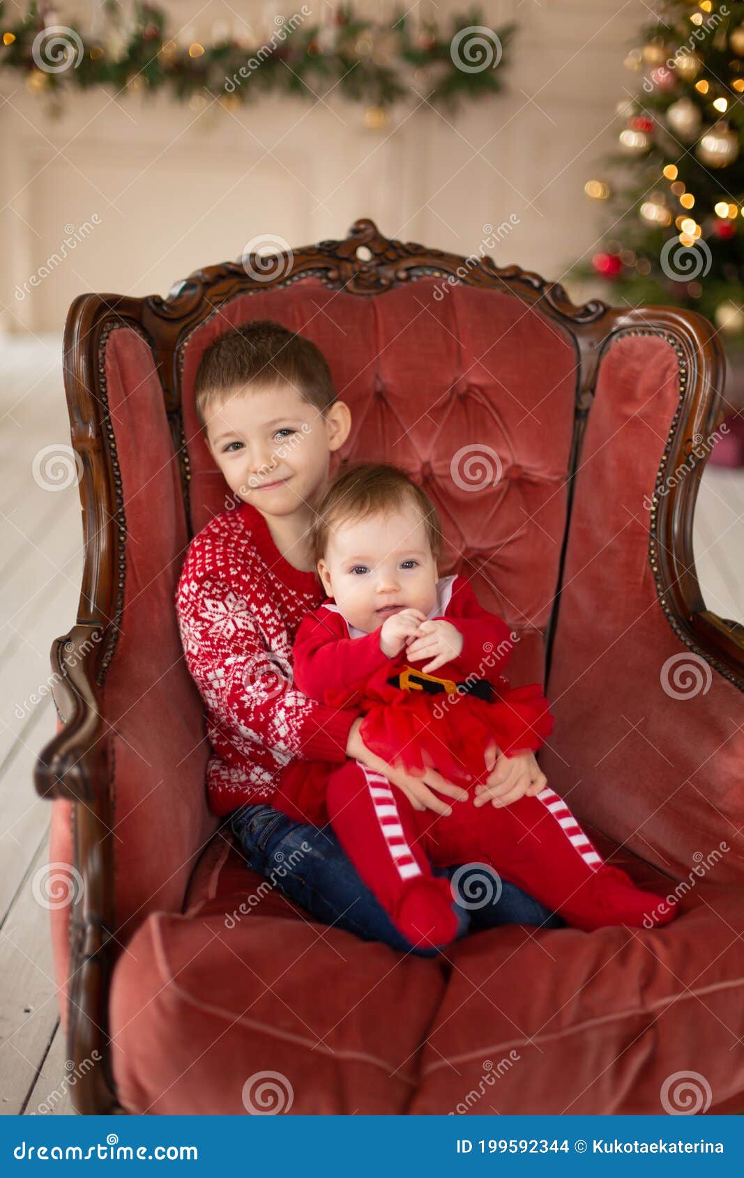 Litttle Brother Hugs His Little Sister In Red Retro Chair Near Christmas Tree Enjoying A Love