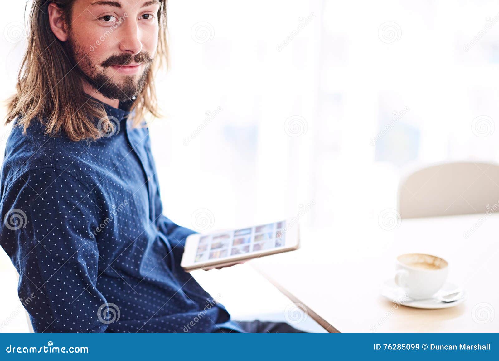 long haired modern man with tablet sitting next to window