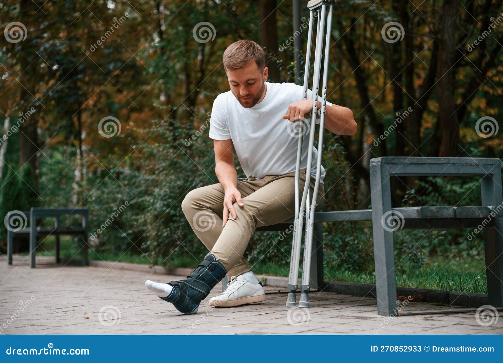 Looking at the Broken Leg. Man with Crutches is in the Park Outdoors